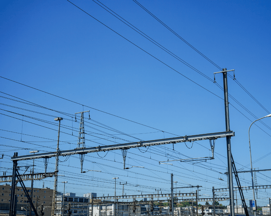 Overhead electric cables and steel support gantries against a clear blue sky, with buildings in the background
